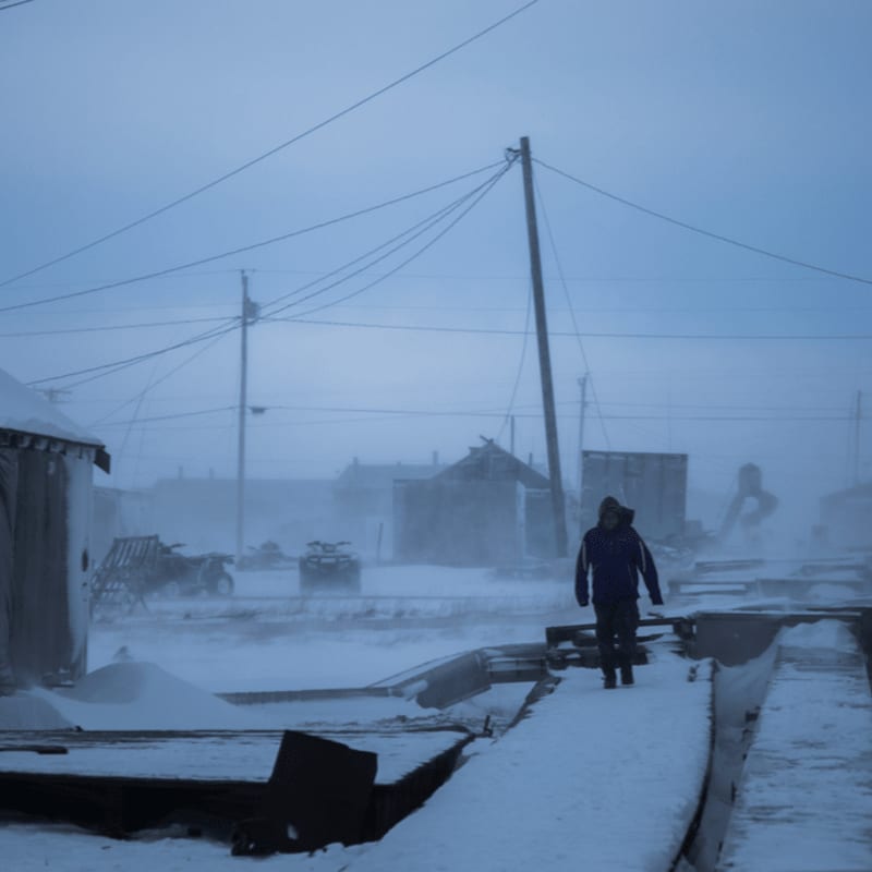 A lone figure walks along a snowy path in a desolate, windswept village under a hazy, overcast sky.