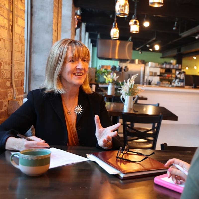 A woman in a black blazer gestures while speaking at a table with a coffee mug and papers in the foreground, while a cafe with hanging lights and a counter is visible in the background.