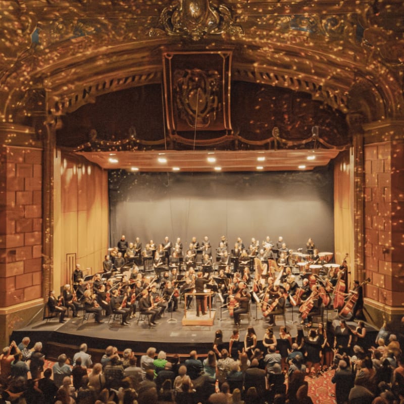 An orchestra performs on a brightly lit stage in a grand, ornate theater filled with an audience.