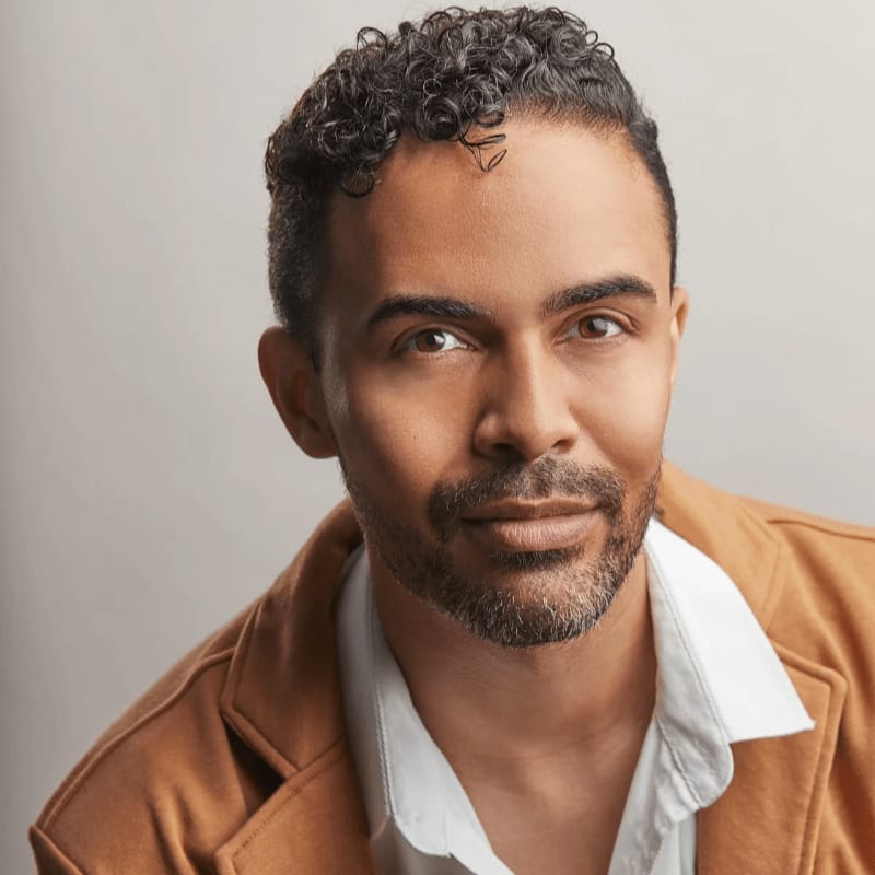 A man with curly dark hair and a light brown jacket over a white shirt looks directly at the viewer against a plain gray background.