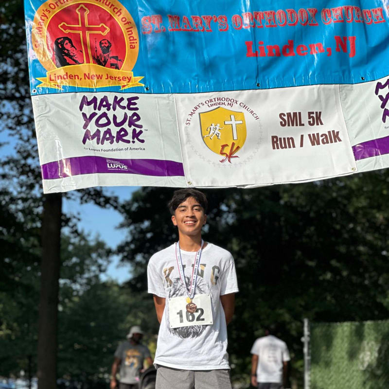 A young man wearing a medal and bib stands in the foreground with a banner for a 5K run/walk in the background.