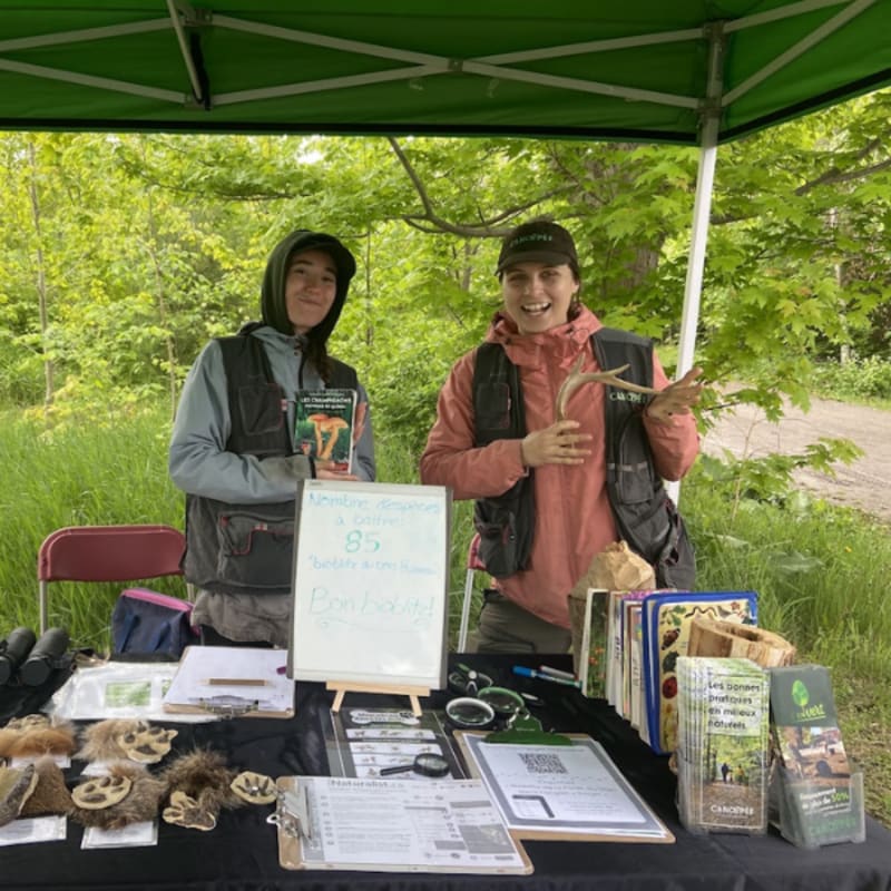 Two people stand behind a table displaying nature-related items, with a wooded area visible in the background.