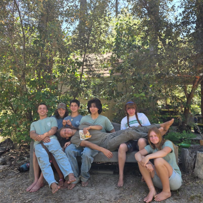 A group of seven young people relax on a bench in front of a wooded area with a building in the background.