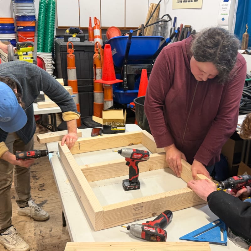 In the foreground, people are assembling a wooden frame with drills, while the background shows a cluttered workshop with tools and safety cones.