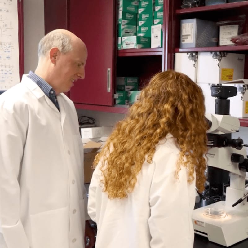 A man in a lab coat looks on as a woman with curly red hair peers into a microscope in a lab filled with shelves of supplies.
