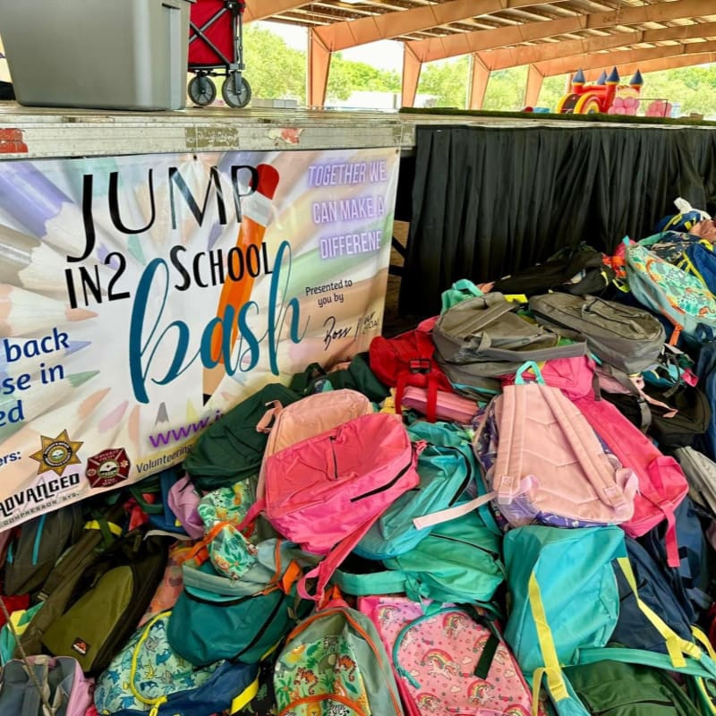 A large pile of colorful backpacks sits in the foreground in front of a banner for a "Jump In2 School Bash," with a bouncy house visible in the background.