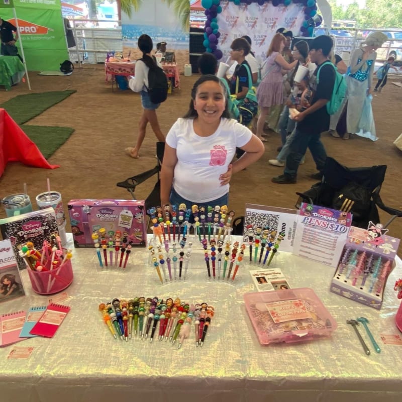 A smiling girl stands behind a table filled with colorful pens and stationery, with a crowd of people and event decorations in the background.