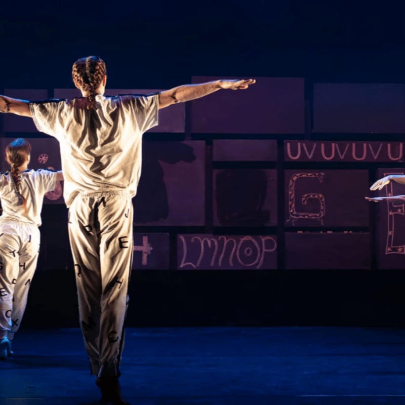 Dancers in white outfits with letters printed on them perform in front of a backdrop of large, stylized letters.