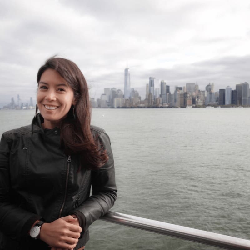 A smiling woman in a black leather jacket stands in the foreground with the New York City skyline visible in the background across the water.