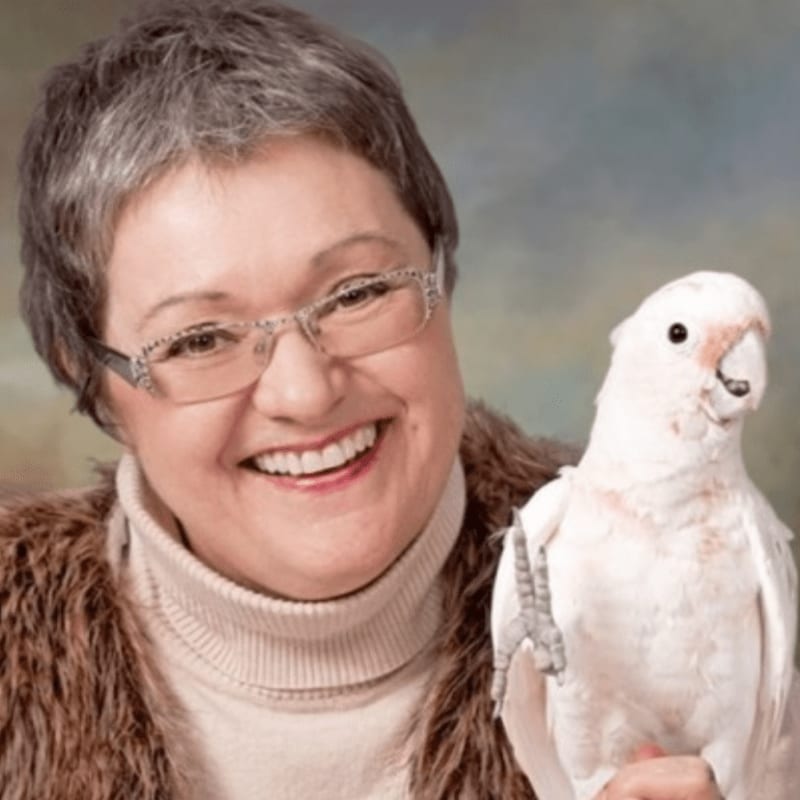 A smiling woman with glasses and a fluffy vest poses with a white cockatoo against a soft, blurred background.