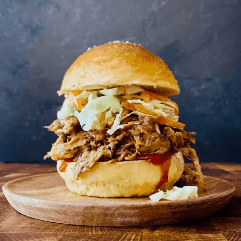 A tall pulled pork sandwich with coleslaw sits on a wooden plate against a dark, textured background.