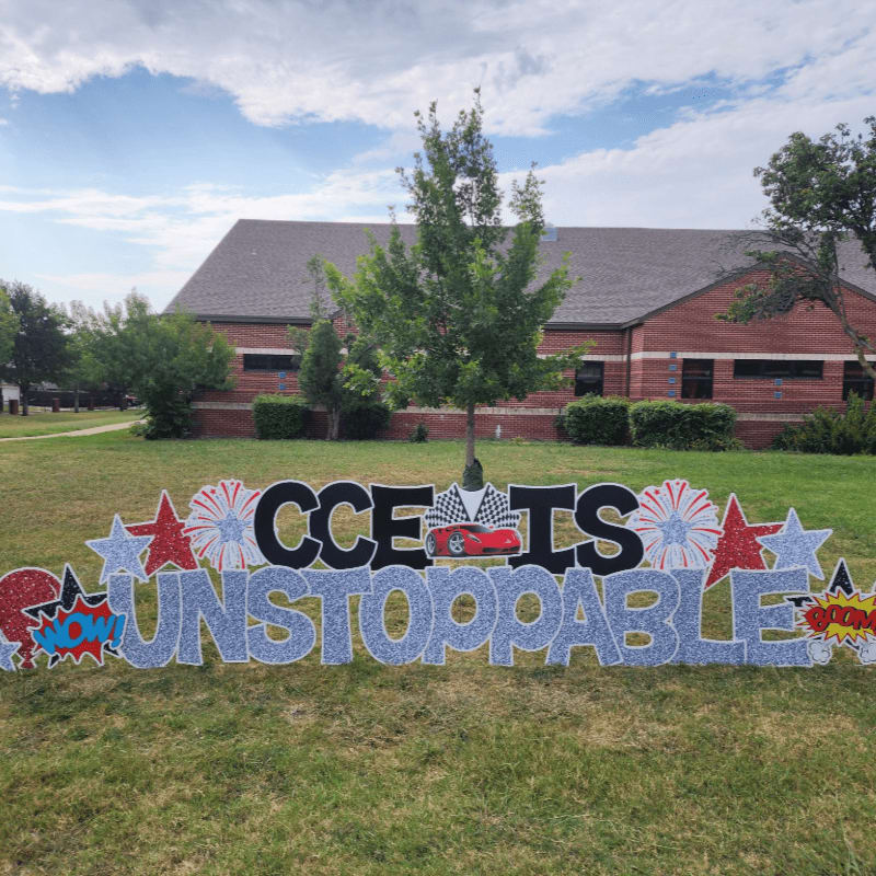 Sparkling blue and red letters spelling "CCE IS UNSTOPPABLE" are displayed on a grassy lawn in front of a brick building with trees.
