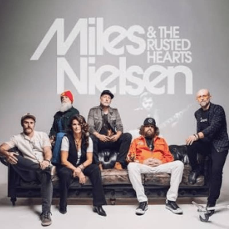 Six musicians, including a woman in the center, are seated on a couch in the foreground, with the band's name "Miles & The Rusted Hearts" displayed in large white lettering against a plain gray background in the upper portion of the image.