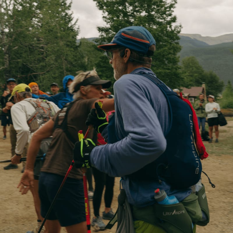 Runners in athletic gear, some with trekking poles, gather in the foreground with a blurred background of trees and distant mountains.