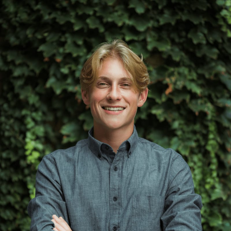 A young man with blond hair smiles with his arms crossed in front of a lush green ivy-covered wall.