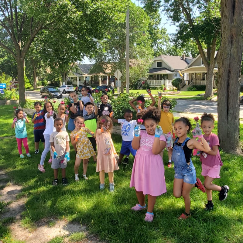 A group of children in the foreground are posing for a photo in a grassy park with houses and trees in the background.