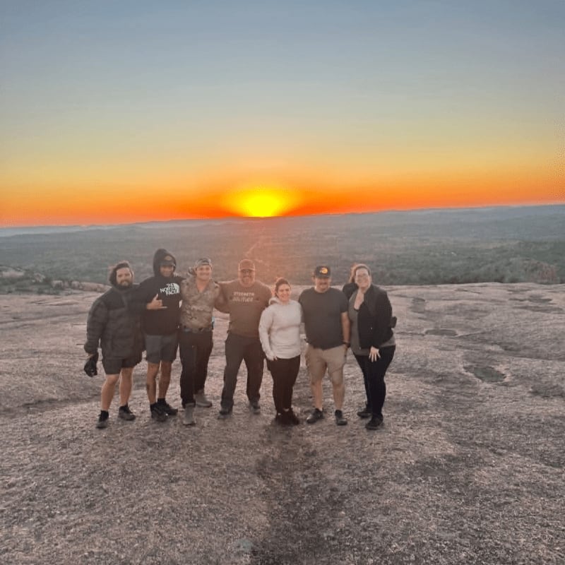 A group of eight people stand on a rocky surface in the foreground, silhouetted against a vibrant orange sunset in the background.
