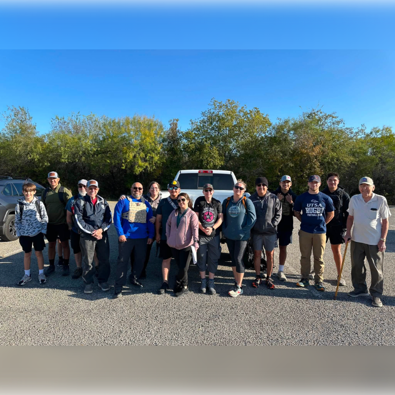 A group of people stand in a parking lot in front of a white truck and a line of trees under a clear blue sky.