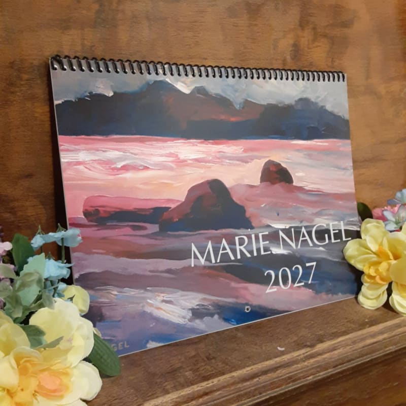 A spiral-bound calendar featuring a painted seascape with rocks in the foreground rests on a wooden surface, adorned with yellow and blue flowers.