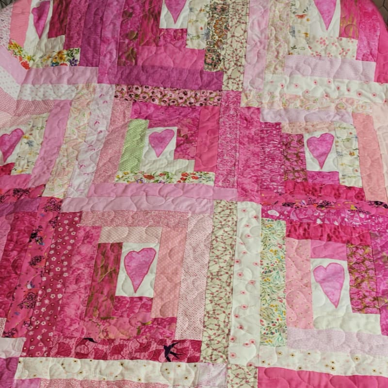 A close-up view of a pink log cabin quilt with a heart in each square fills the frame, with no background visible.