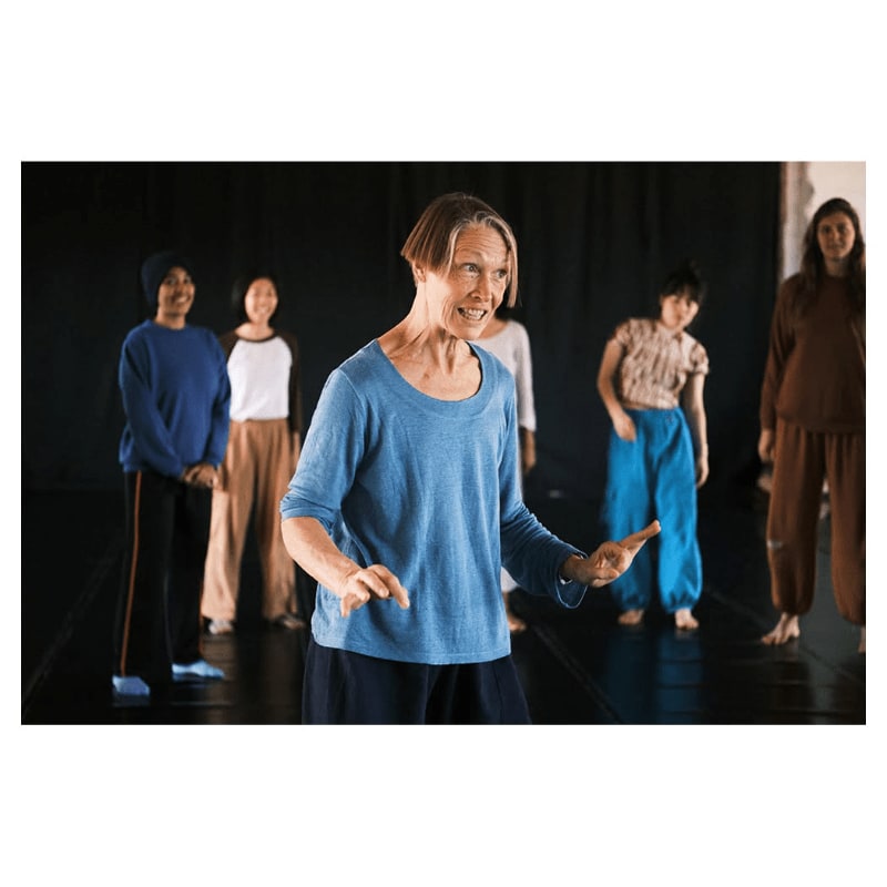 A woman in a black t-shirt and grey pants smiles as she dances, with two blurred figures in the background against a light sky.