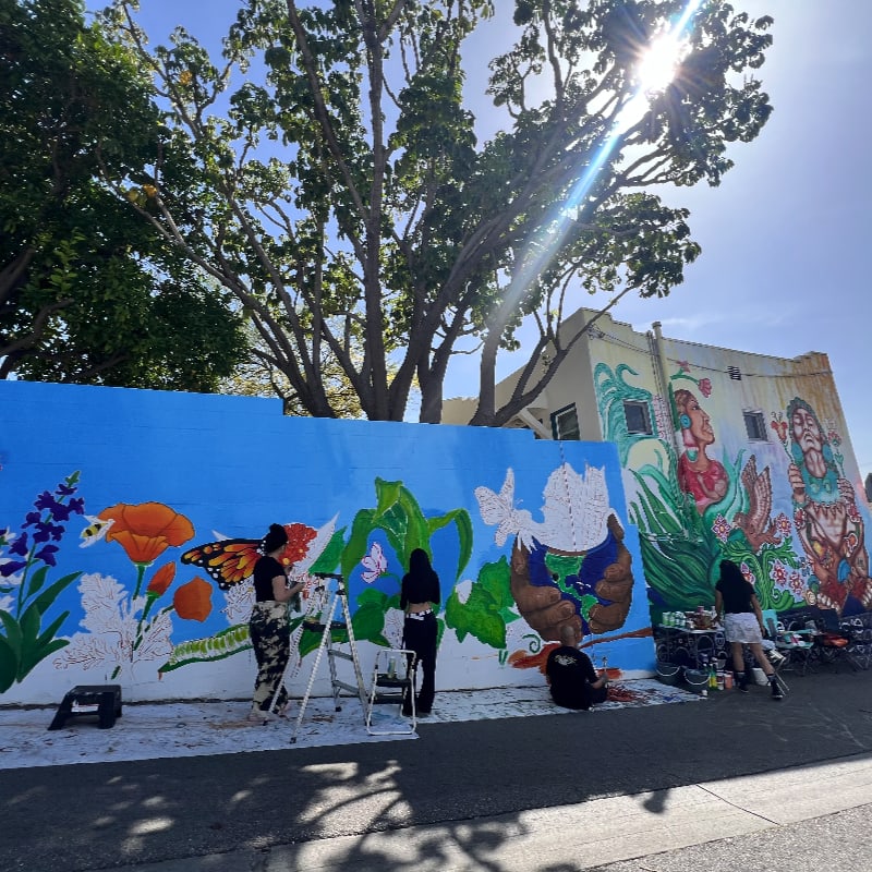 In the foreground, artists paint a vibrant mural of flowers and butterflies on a blue wall, while in the background, a sunlit building and trees frame the scene.