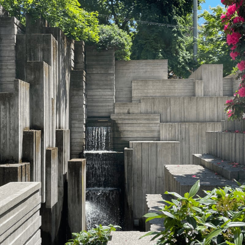 In the foreground, lush green foliage and a gravel path lead to a striking concrete waterfall structure, while the background reveals a vibrant pink flowering bush against a backdrop of green trees and a clear blue sky.