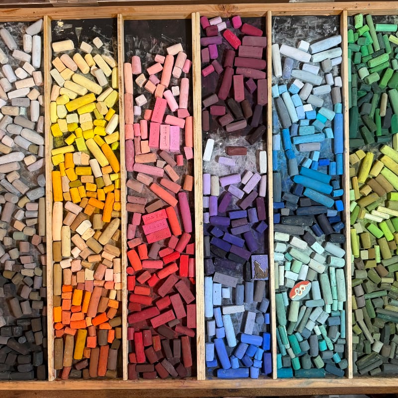 A wooden tray is filled with rows of colorful pastel chalk sticks, ranging from grays and yellows to reds, purples, blues, and greens.