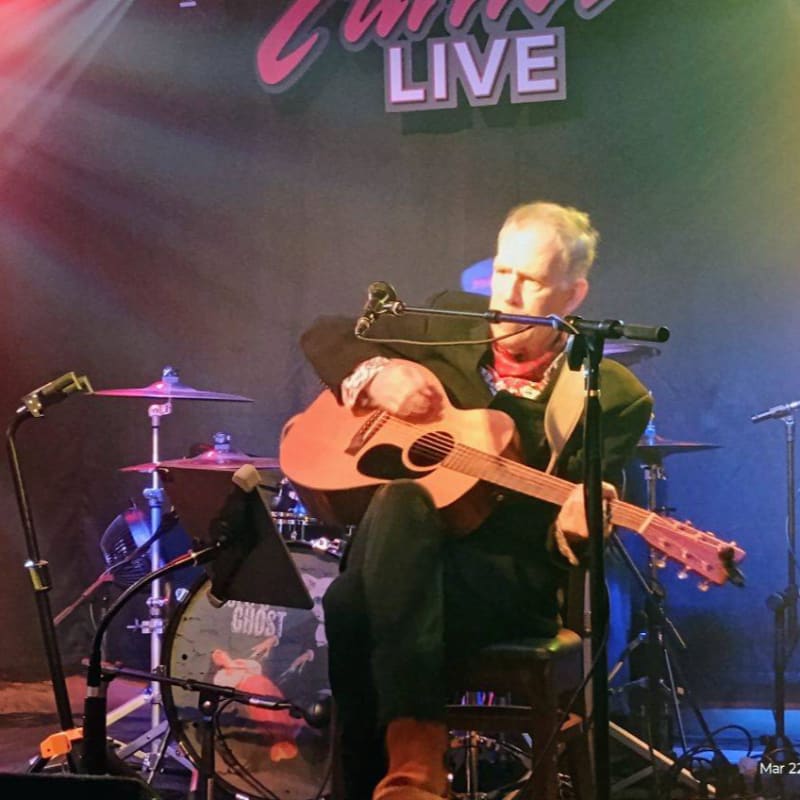 A man plays an acoustic guitar on stage in front of a drum set, with a "LIVE" sign illuminated in the background.