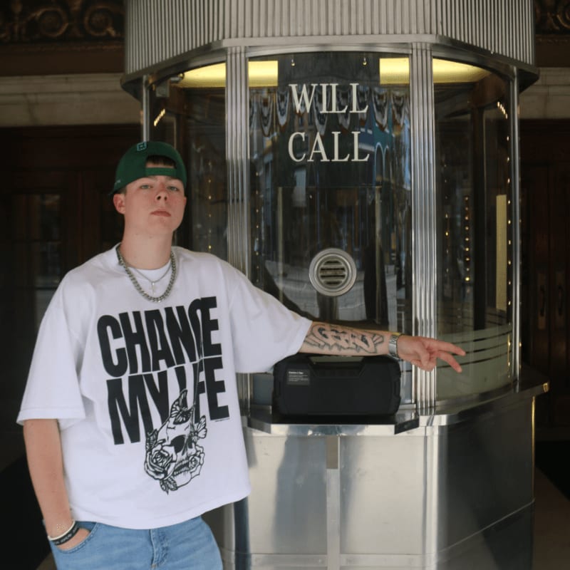 A young person in a white t-shirt and green hat stands in front of a "Will Call" booth with a metallic, art deco design.