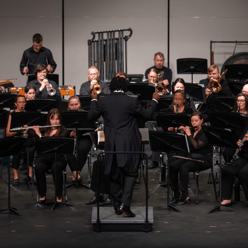 A conductor directs a band of musicians playing various instruments in front of a plain stage backdrop.