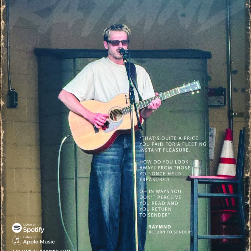 A man in sunglasses and a t-shirt plays an acoustic guitar and sings into a microphone in front of a large metal door, with a chair and traffic cone visible to the right.