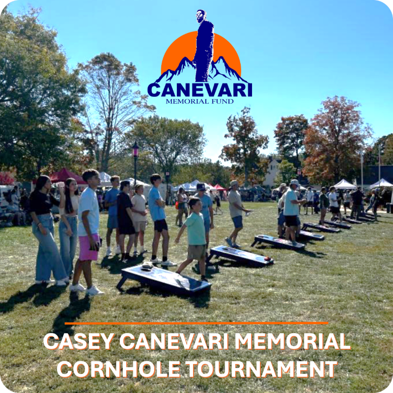 People play cornhole on a grassy field in front of tents and trees under a bright blue sky with a memorial fund logo.