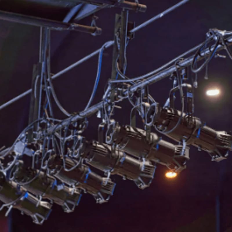 Stage lights hang from a metal grid in the foreground, with a dark, blurred background suggesting a performance space.
