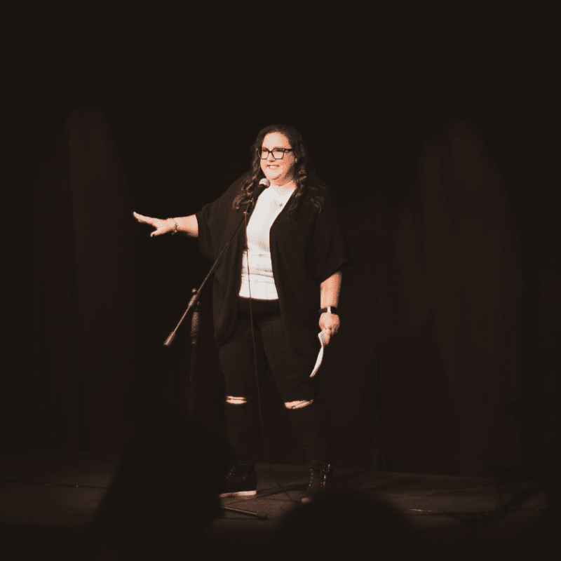 A woman in ripped jeans and a black cardigan gestures with her left hand while holding a paper in her right, standing on a dimly lit stage with a dark curtain behind her.