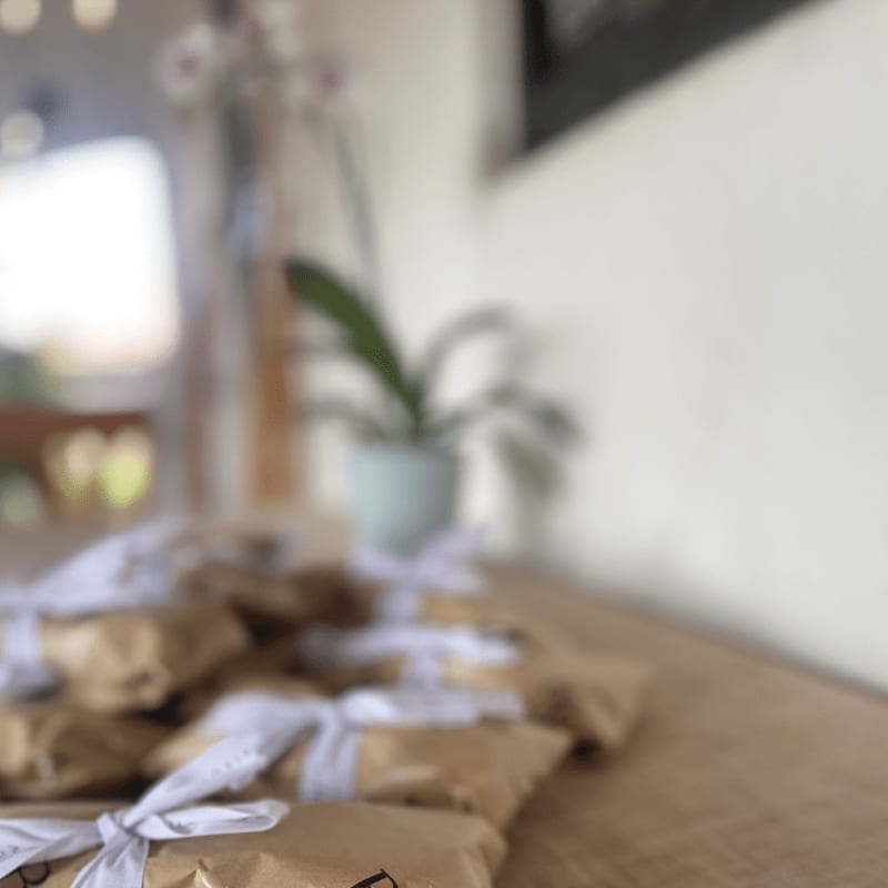 Brown paper-wrapped gifts with white ribbons are arranged on a wooden surface in the foreground, with a blurred potted plant and a bright window in the background.