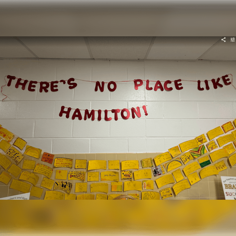Red glitter letters spelling "THERE'S NO PLACE LIKE HAMILTON!" hang above a wall decorated with yellow paper squares, some with drawings and writing, against a white brick background.