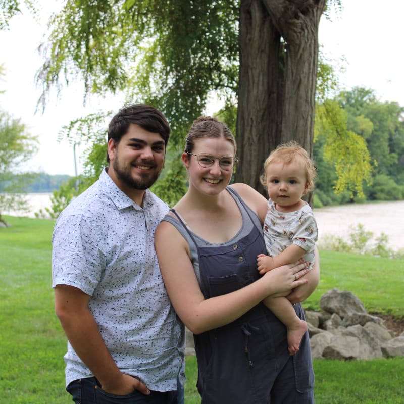A smiling family of three, with a man and woman holding a baby, stand in a grassy park with a river and trees in the background.