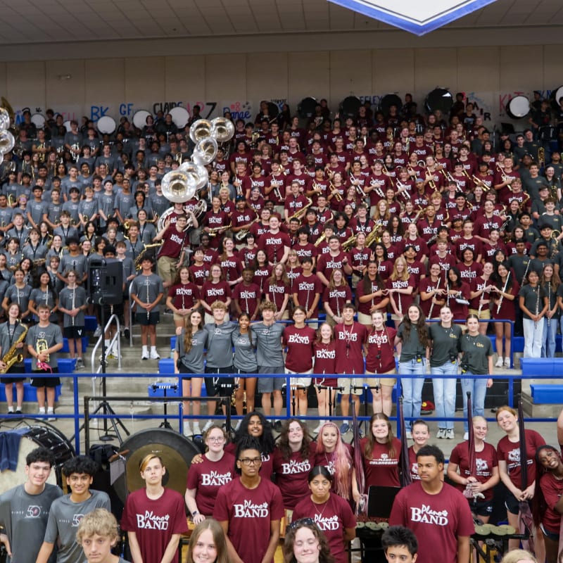 A large marching band in maroon shirts and grey pants poses for a photo in a gymnasium, with rows of students and instruments filling the frame from the foreground to the bleachers in the background.