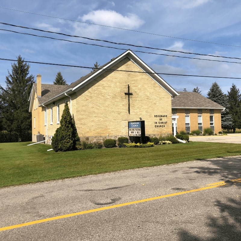 A yellow brick church with a cross on its facade stands on a grassy lawn in front of a line of trees under a partly cloudy sky, with a road in the foreground.