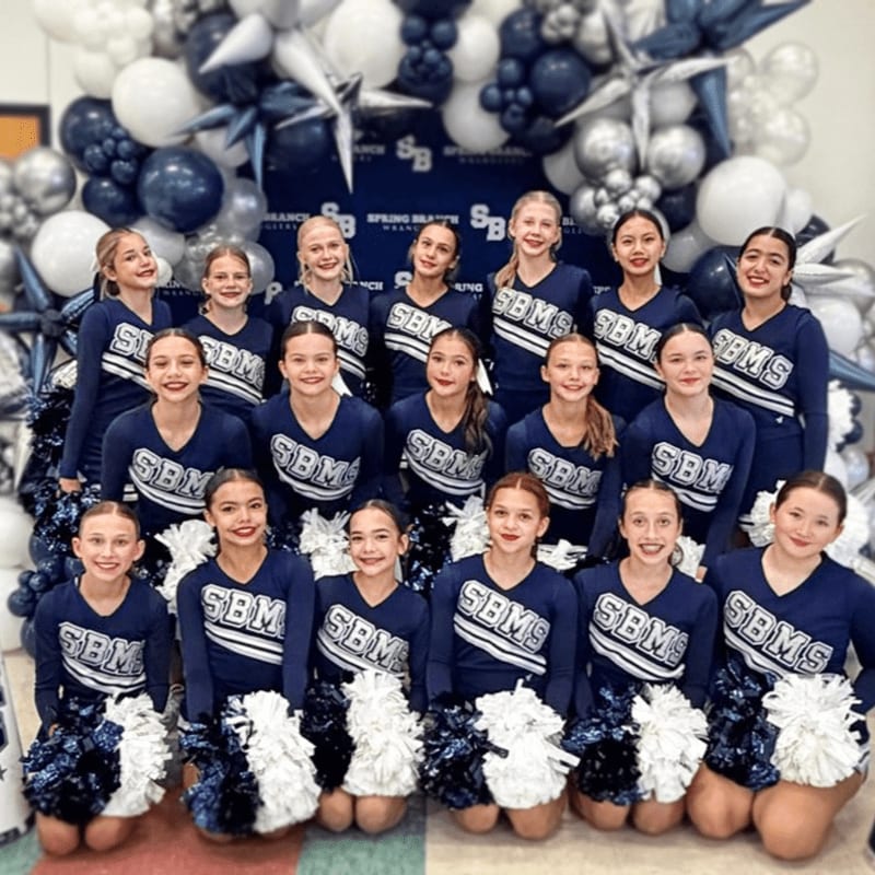 A group of young cheerleaders in navy blue uniforms with "SBM" on them pose in front of a balloon arch.