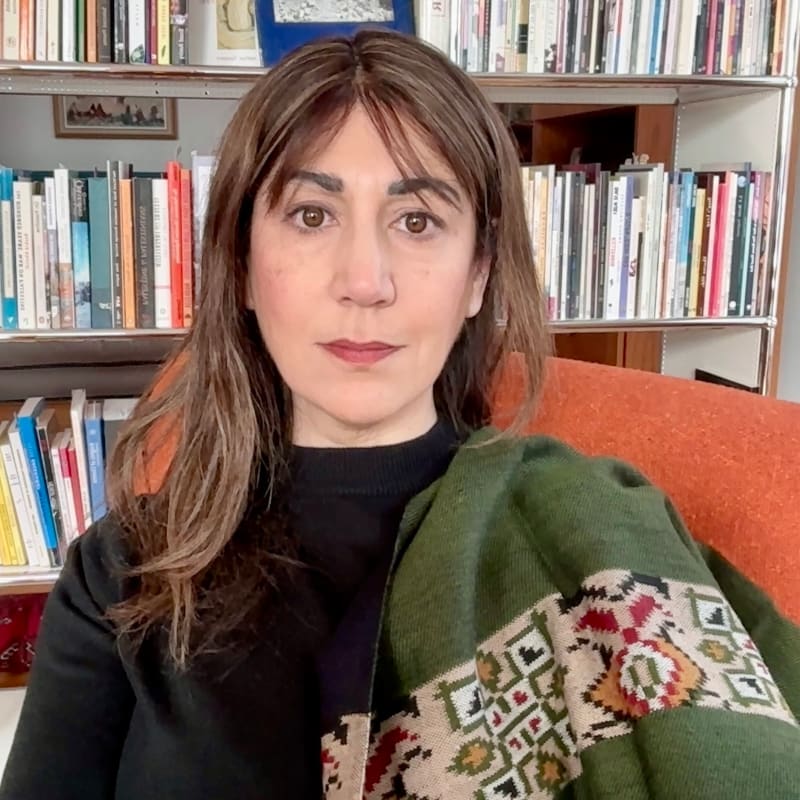 A woman with brown hair and a green patterned shawl sits in front of a bookshelf filled with books.