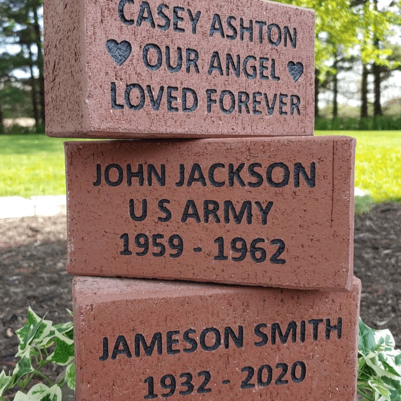 Three engraved memorial bricks are stacked in the foreground, with a blurred background of green grass and trees.
