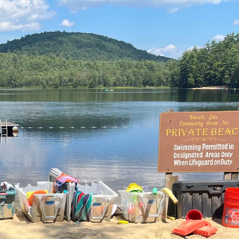 Beach gear sits on the sand in the foreground, with a "Private Beach" sign and a calm lake reflecting a tree-covered hill in the background.