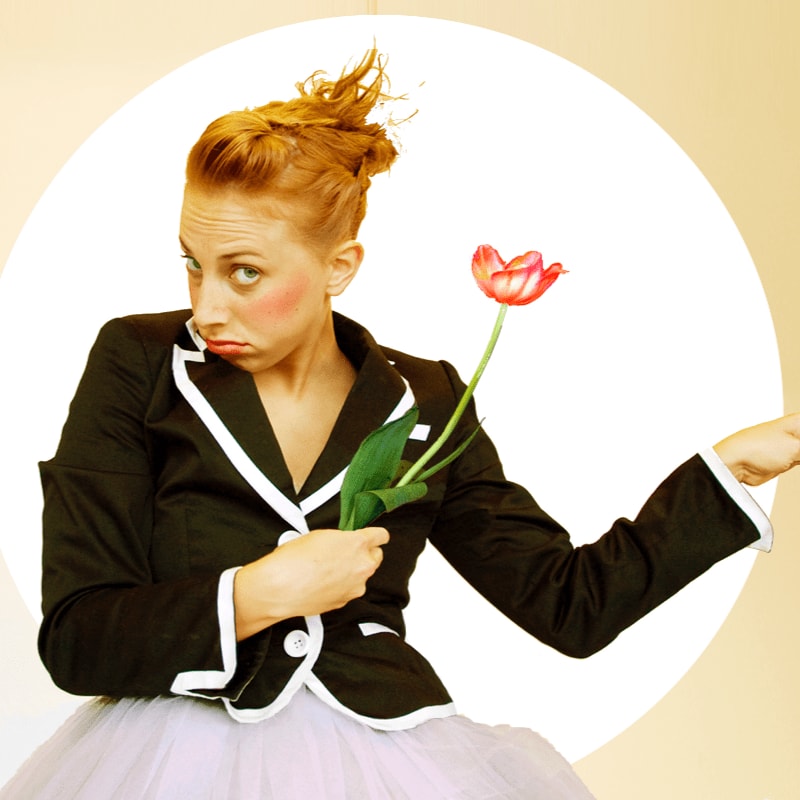 A woman with red hair and a pouty expression holds a single red tulip in the foreground, while a soft yellow circle forms the background.