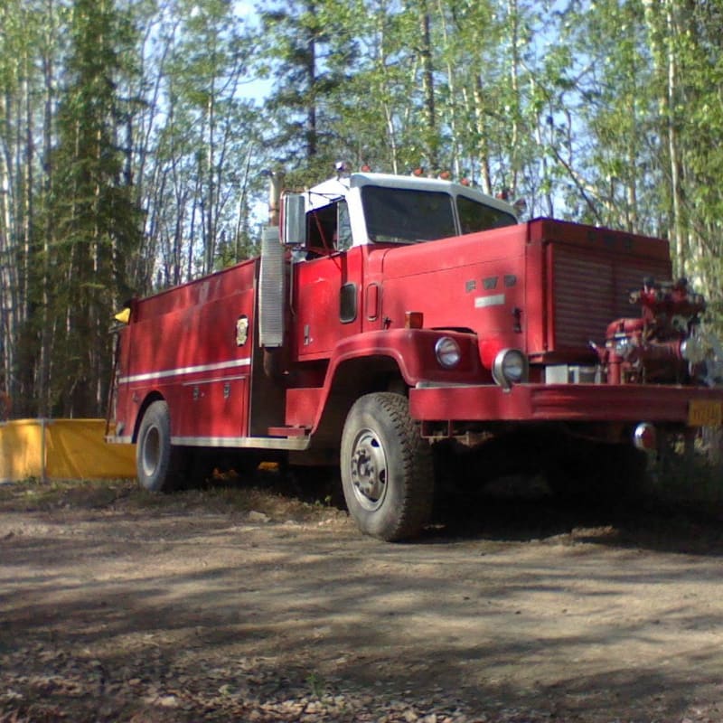 A red fire truck sits on a dirt road in front of a dense forest.