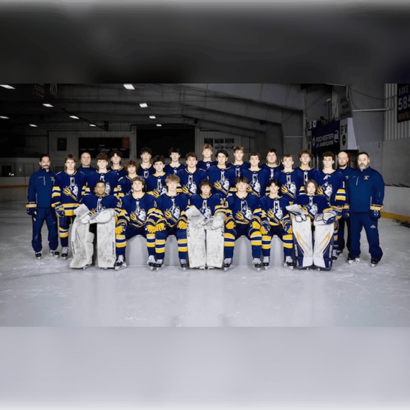A hockey team in blue and yellow uniforms poses on the ice in front of a rink with bleachers in the background.