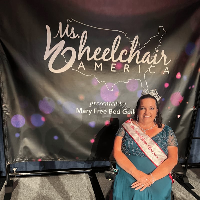 A woman in a teal gown and a sash sits in a wheelchair in front of a "Ms. Wheelchair America" banner.