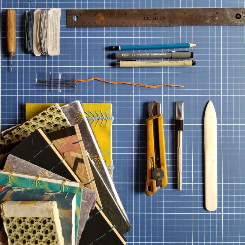 In the foreground, a collection of handmade books and bookbinding tools are laid out, while the background features a blue grid mat and a metal ruler.