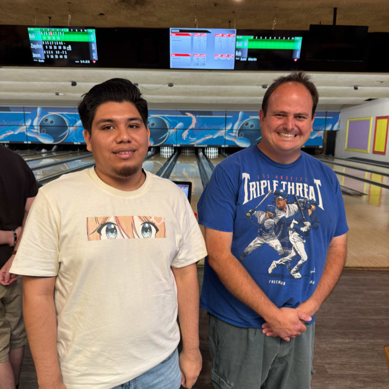 Two men stand in the foreground of a bowling alley, with bowling lanes and scoreboards visible in the background.